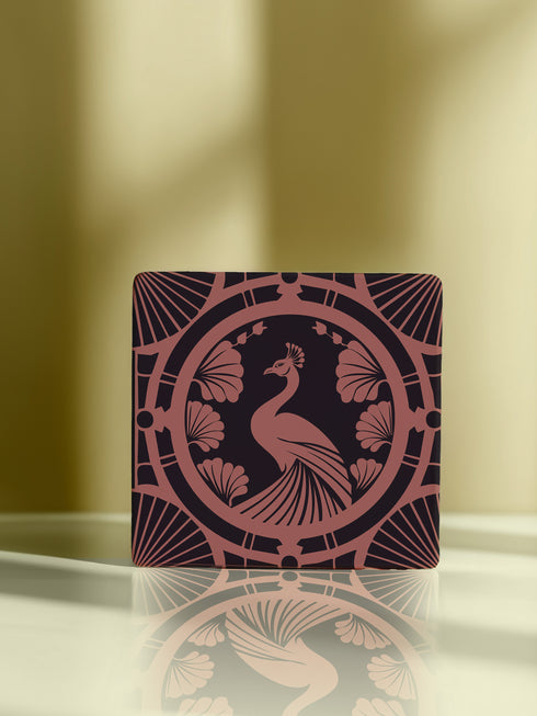 Square ceramic coaster featuring a detailed Art Deco peacock design in copper tones, surrounded by shell motifs and geometric fan patterns on a dark charcoal background, with protective cork base.